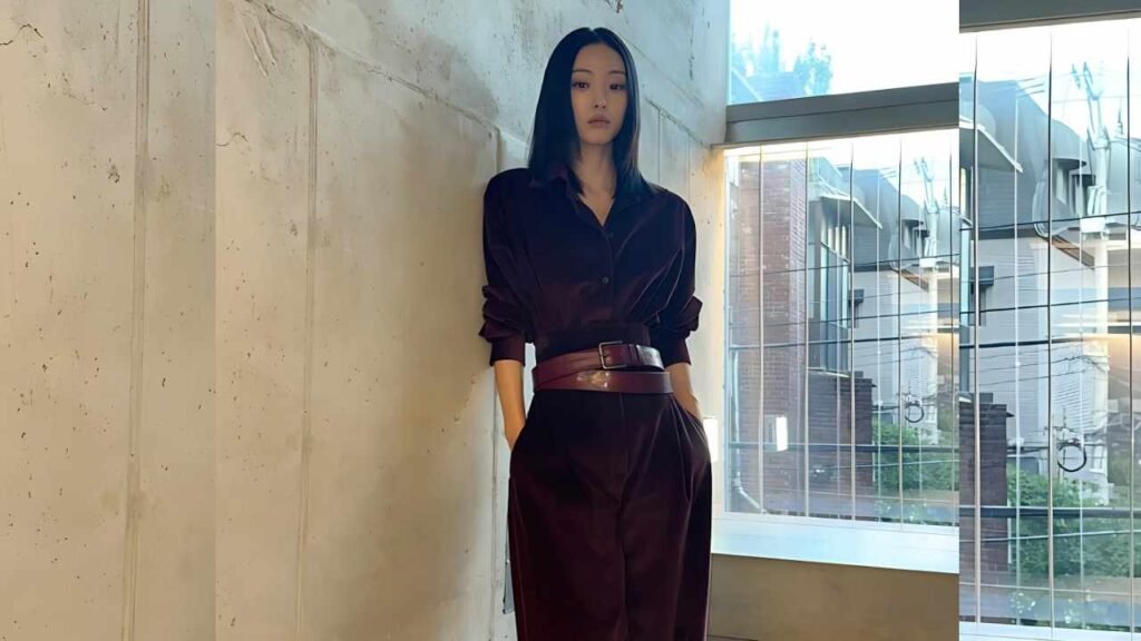 Jeon So-nee in a burgundy velvet suit with a wide belt stands with hands in pockets against a concrete wall beside a glass atrium, cityscape beyond.