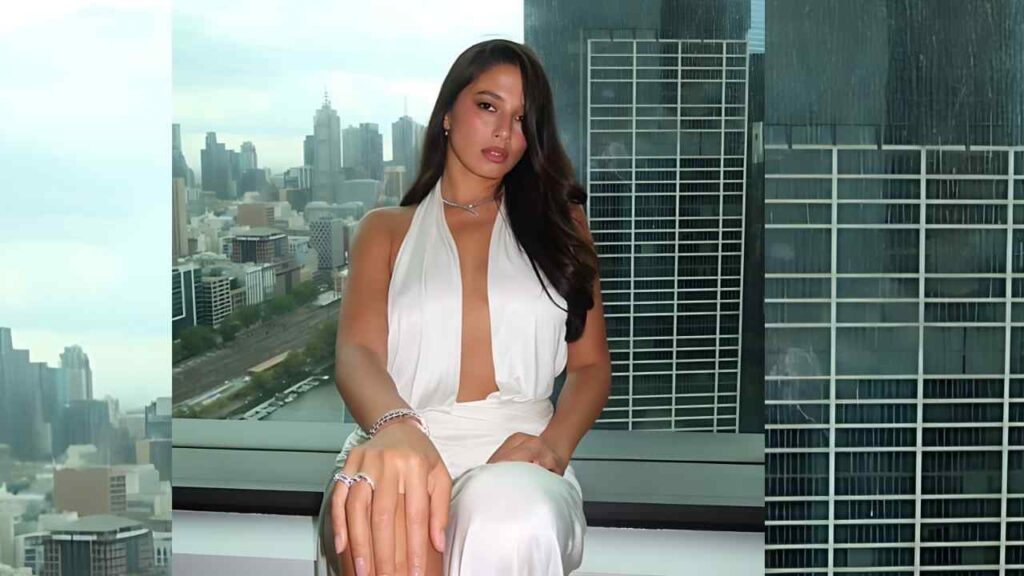 Jessica Gomes in a white halter dress sits on a windowsill, city skyline visible behind; long dark hair and silver jewelry.