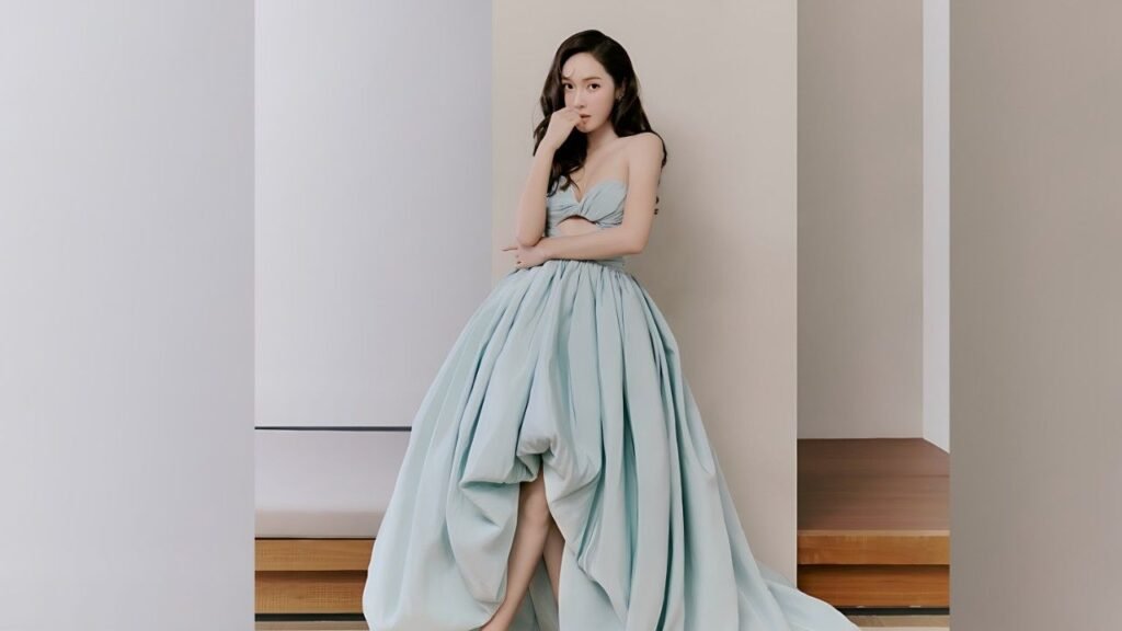 Jessica Jung in a pale blue strapless ball gown stands against pastel walls, one hand near her mouth, gaze soft.