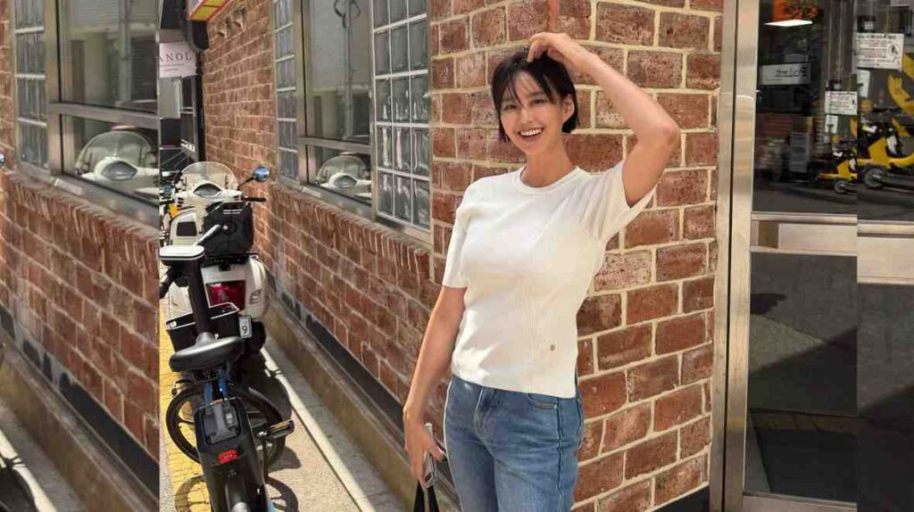 Ji So-yeon (지소연) in a white T-shirt and blue jeans stands beside a parked scooter along a brick street, hand on her head.