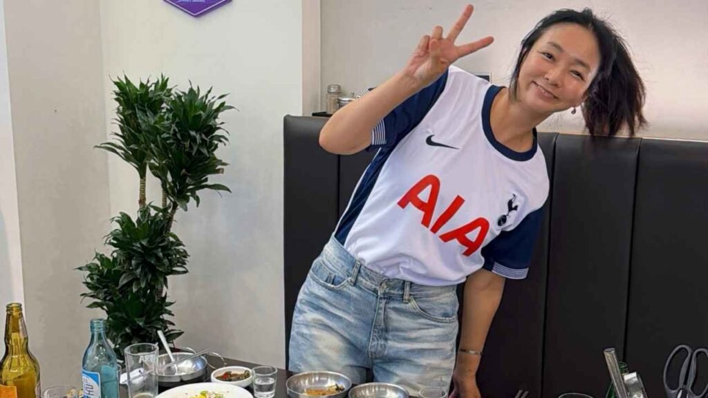 Jin Jae-young in white and navy jersey with AIA letters and jeans poses peace-sign beside a plant at a booth with dishes.