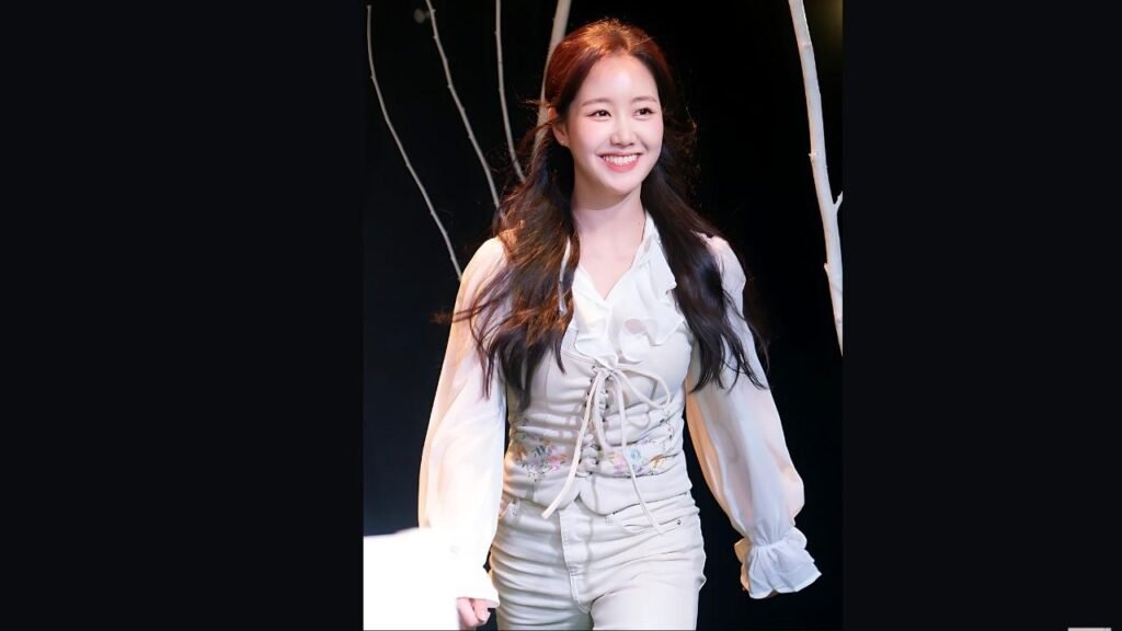 Jin Ji-hee in a white ruffled blouse and fitted white pants walks on a dark stage, with pale tree props behind her.