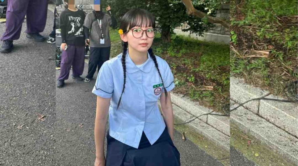 Jin Ki-joo with round glasses and two braids in a light blue uniform top with a badge, standing on a paved path outdoors.