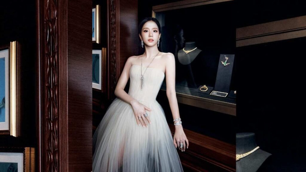 Jisoo in a strapless pale ombré tulle gown poses beside a jewelry display in a dark wood-paneled room.