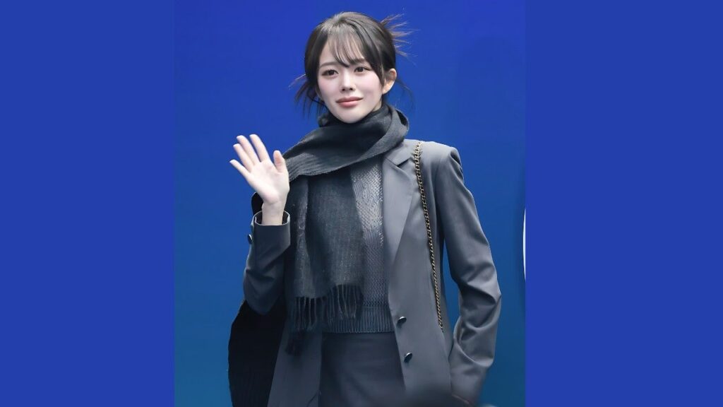 Jung Ji-so in a gray suit and scarf waves hello, carrying a chain-strap bag, against a blue gradient backdrop.