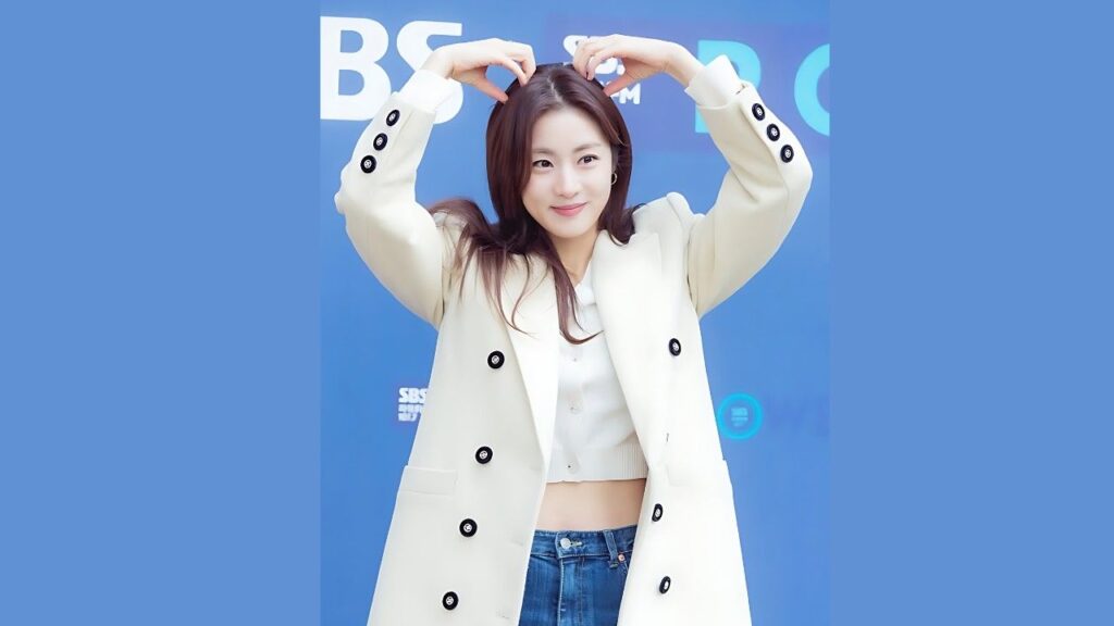 Kang Jung-hwa in a cream double-breasted coat over a white crop top and jeans forms a heart with her arms against blue backdrop.