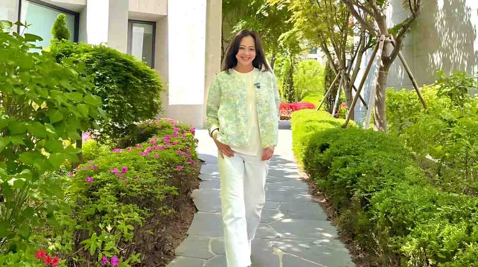 Kang Sung-yeon walks a stone path, flanked by green hedges and pink flowers, wearing a pale green jacket and white pants.