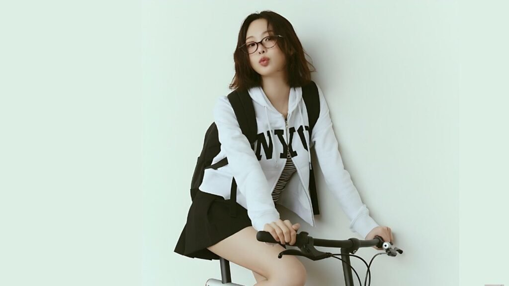 Keum Sae-rok with glasses sits astride a bike, white hoodie with NY lettering, black backpack, against a pale mint wall.