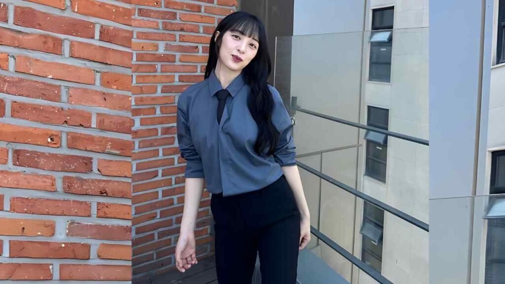 Kim Bo-ra with long black hair in a blue-gray shirt and black tie, leaning on a brick wall beside a glass-railed balcony.
