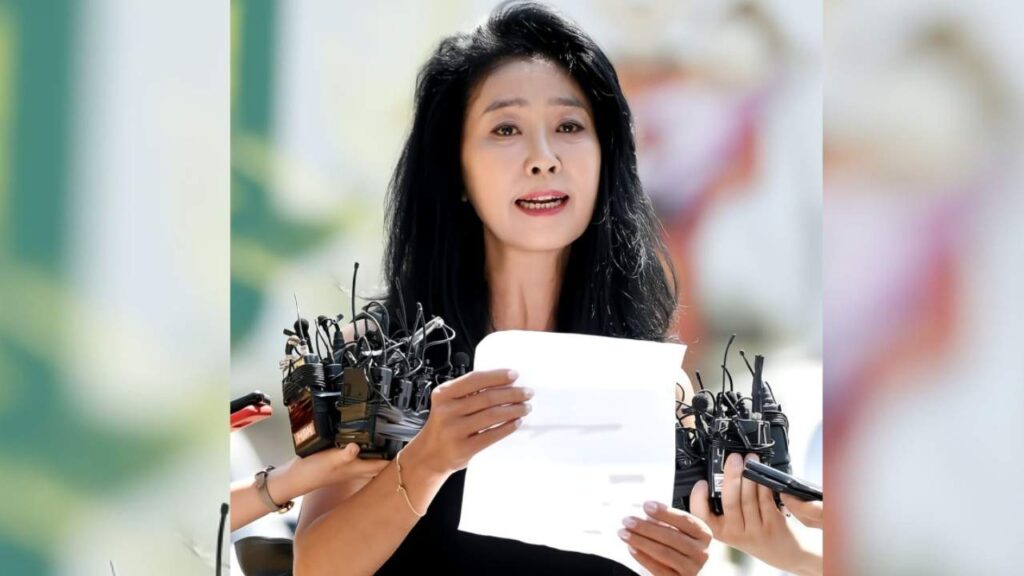 Kim Boo-sun (김부선) in black top speaks to press, holding a sheet of paper while numerous lavalier microphones crowd around her outdoors.