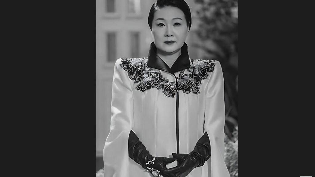 Kim Hae-sook in a white satin cape with black floral embroidery, with gloves and hands clasped.