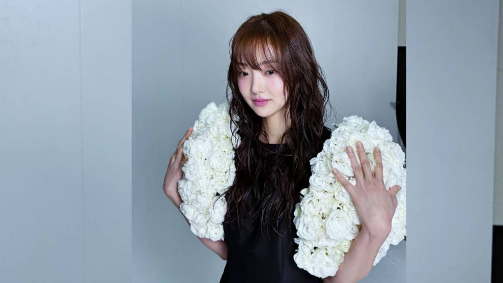 Kim Hye-jun with damp, wavy hair in a black top holds two large white rose bouquets against pale panels, looking thoughtful.