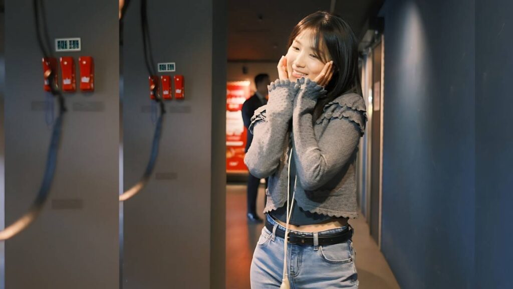 Kim Hye-yoon in a cardigan and denim jeans stands in a blue hallway, hands on her cheeks; fire-alarm boxes line on left.