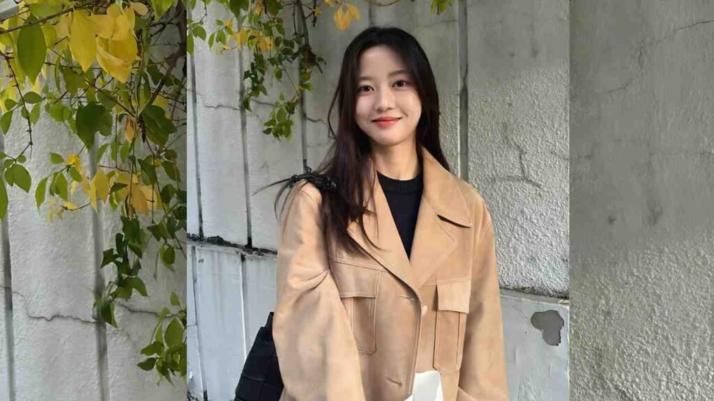 Kim Hyun-soo (김현수) with long dark hair wearing a tan oversized jacket over a black top standing against gray concrete wall, smiling.