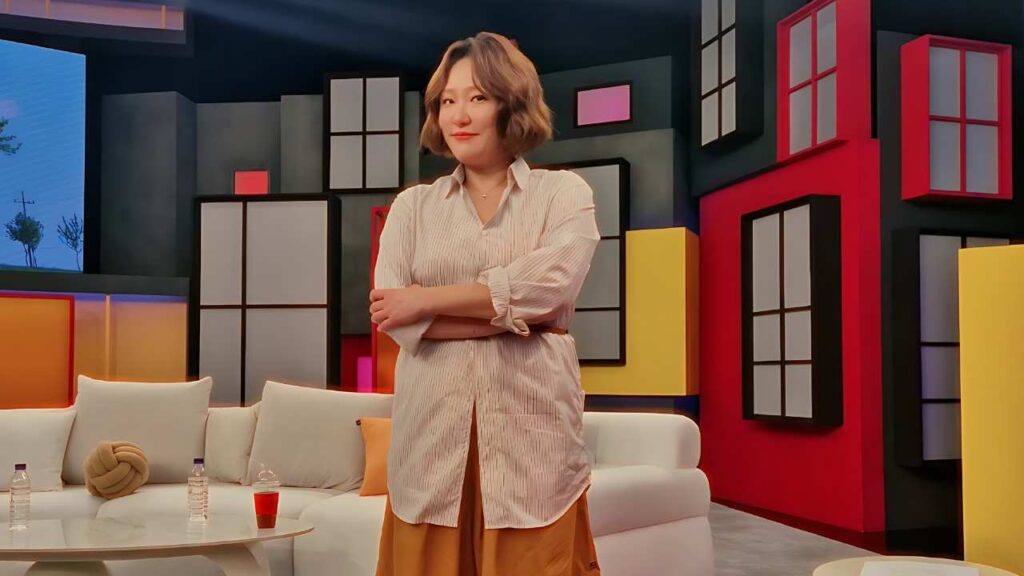 Kim Hyun-sook (김현숙) with short brown hair stands with arms crossed in a vibrant geometric studio set near a white sofa and table.