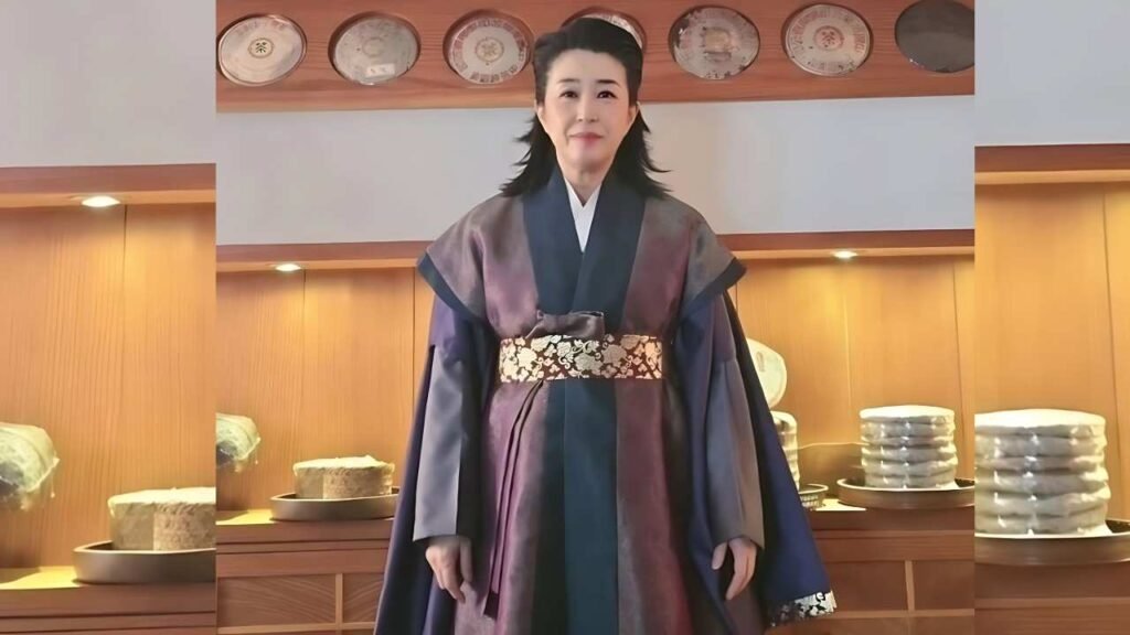Kim Mi-kyung in a purple-to-navy traditional robe with a gold floral obi stands in a warmly lit pastry shop, cakes on wooden shelves behind.