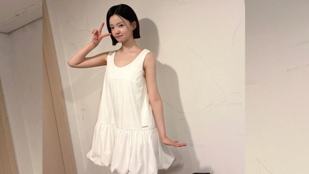 Kim Si-a with a short black bob in a white sleeveless dress poses playfully, peace sign near her head, against a pale wall.