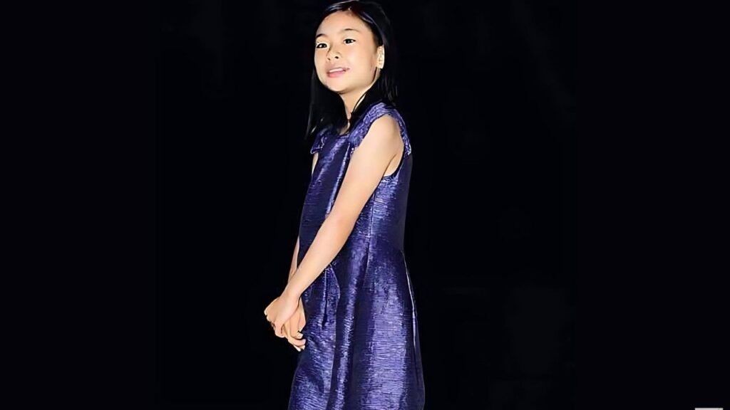 Kim Su-an (김수안) in a shiny purple sleeveless dress stands sideways against a dark background, hands clasped, softly smiling.