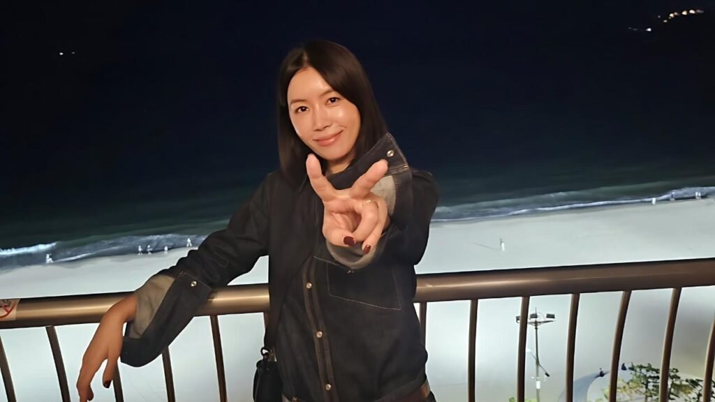 Kim Yoo-mi in a dark denim jacket leans on a railing, flashing a peace sign with a smile as waves roll behind.