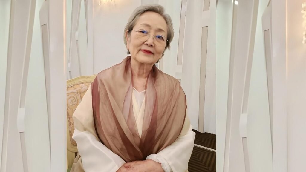 Kim Young-ok with short gray hair and glasses sits in a cream chair, wearing a white blouse and a sheer brown scarf.