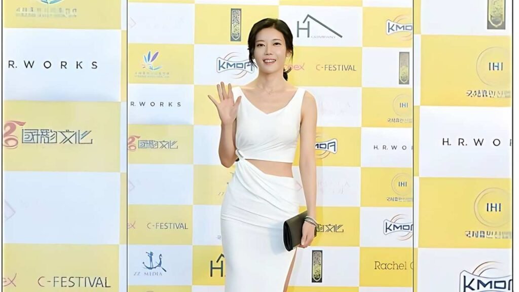Ko Won (고원) in a white crop top and long skirt waves at a yellow-and-white checkered backdrop, holding a black clutch.