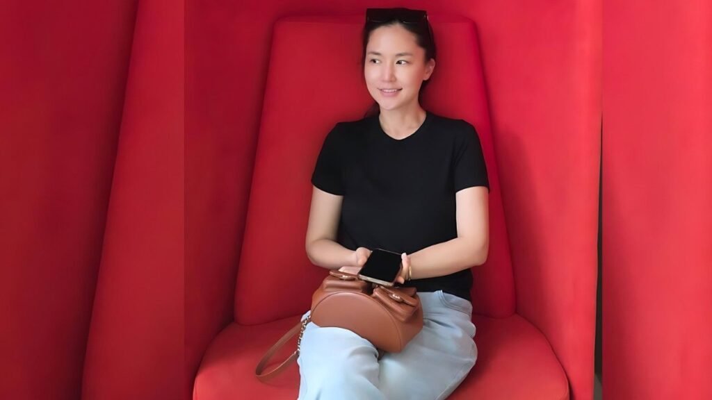 Kong Hyun-joo sits in a red booth, wearing a black top and light jeans, holding a phone with a brown handbag on her lap.