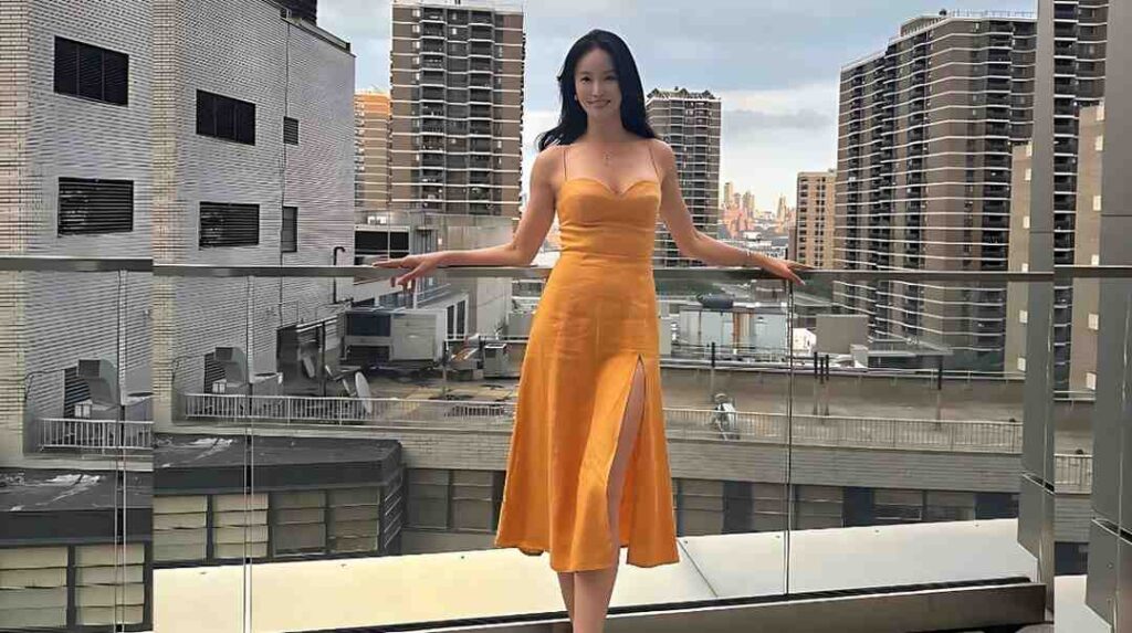 Lee Eon-jeong (이언정) in a bright orange strappy midi dress with a thigh slit leans on a glass balcony railing, with city high-rises behind.