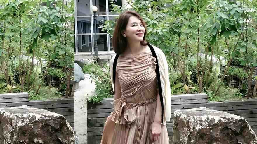 Lee Il-hwa in a taupe ruched, pleated dress with a belt stands in a lush garden beside wooden planters and rocks.