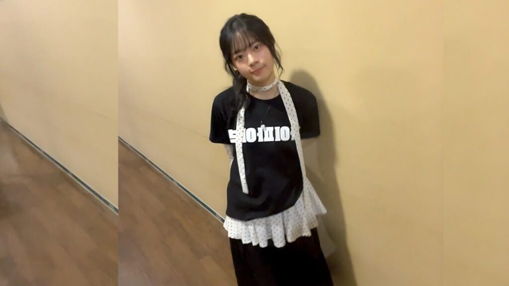 Lee Jae-in with dark hair and bangs leans against a pale beige hallway wall, wearing a black tee with white print, a white polka-dot scarf, and a layered white ruffled skirt over black pants.