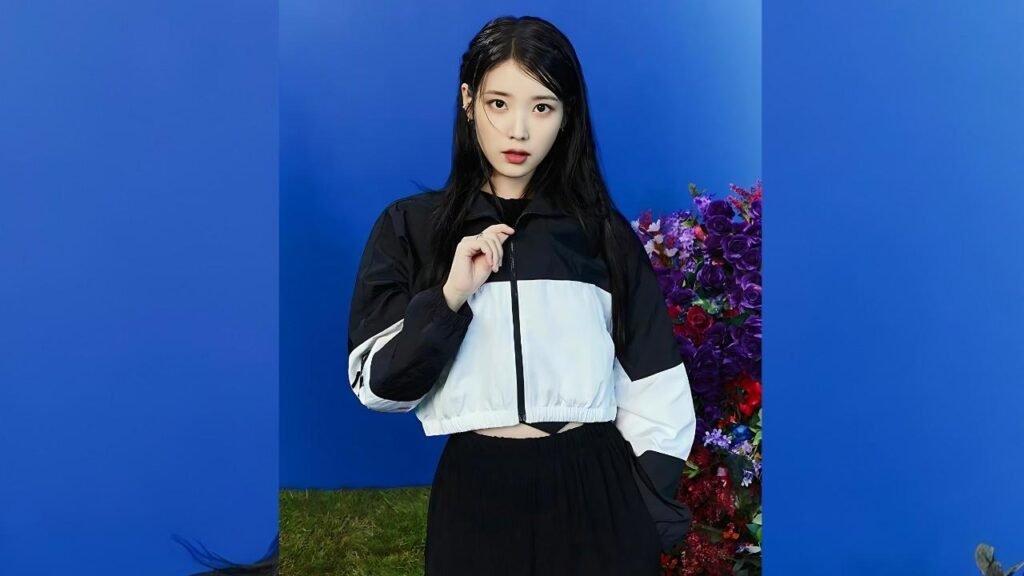 Lee Ji-eun with long black hair, wearing a black-and-white cropped jacket, poses before blue panels with flowers.