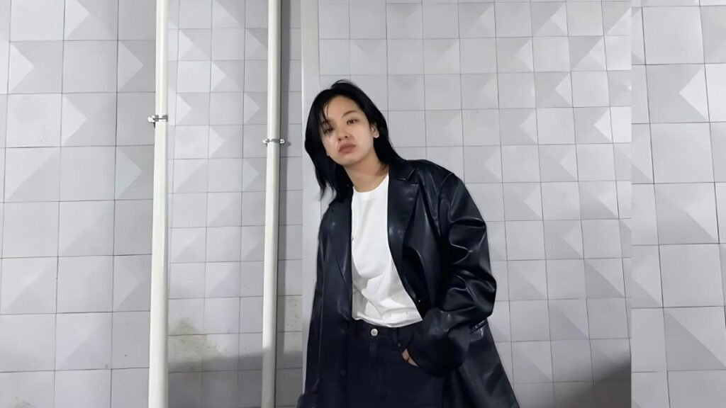 Lee Joo-young in a black leather coat over a white tee leans slightly against a pale tiled wall with pipes, looking at the camera.