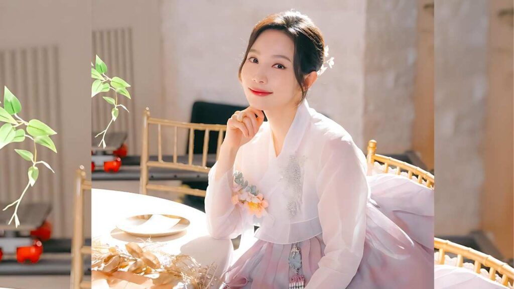 Lee Min-young (이민영) in pale pink hanfu sits at a white table, hand under chin, floral embroidery, bun with a white flower.