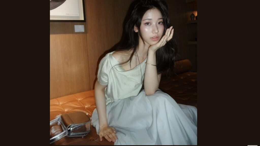 Lee Re sits on orange tufted sofa in a pale blue dress, resting her head on her hand; a silver purse lies nearby.