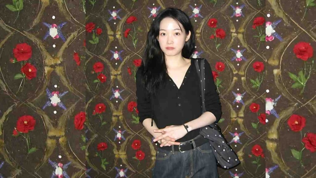 Lee Yoo-mi with long black hair in a black top and jeans, hands clasped, stands before floral wallpaper featuring red roses.