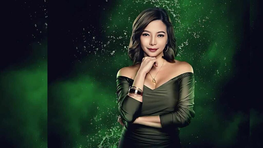 Maui Taylor in off-shoulder olive-green dress with gold jewelry and hoops, chin on her hand against emerald backdrop.