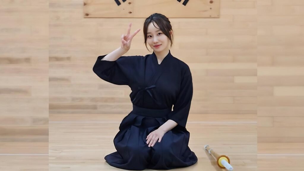 Moon Ye-won in a navy kimono sits kneeling on a light wooden dojo floor, flashing a peace sign with her right hand and smiling, baton at her side.