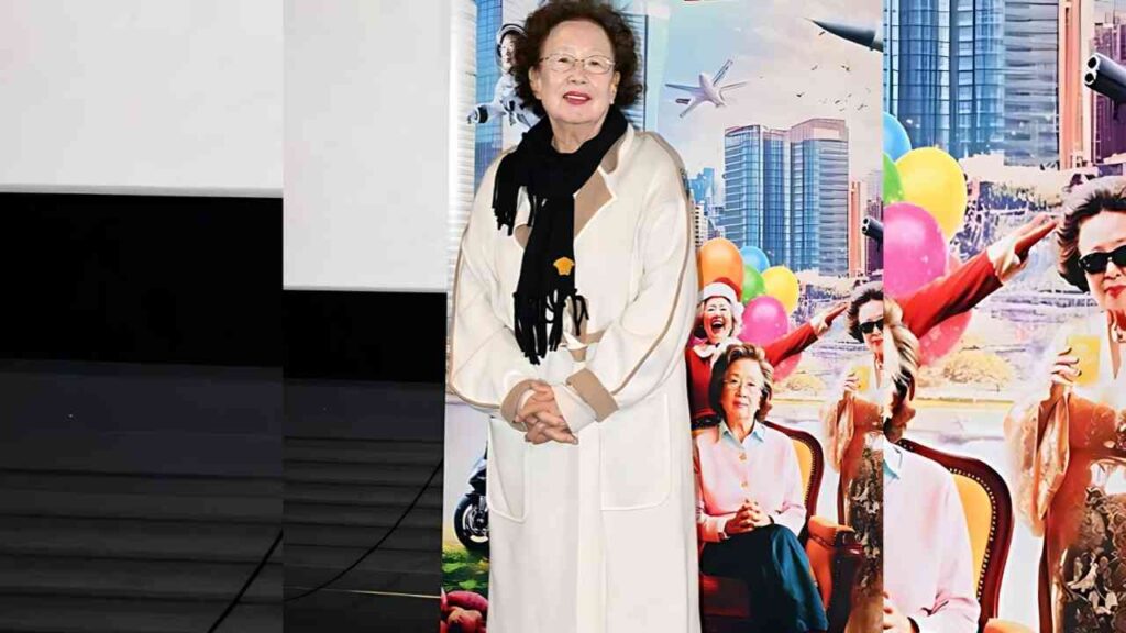 Na Moon-hee with short curly hair and glasses, wearing a cream coat and black scarf, stands with clasped hands before a colorful backdrop featuring balloons and city skyscrapers.