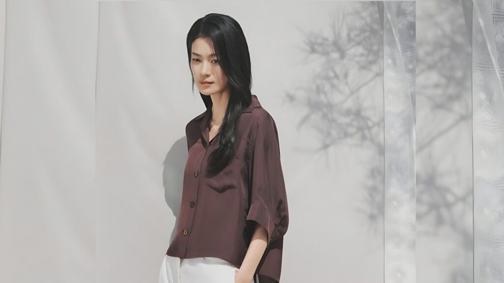 Ok Ja-yeon with dark hair wears a loose maroon button-down shirt and white pants, standing against white wall with tree shadows.