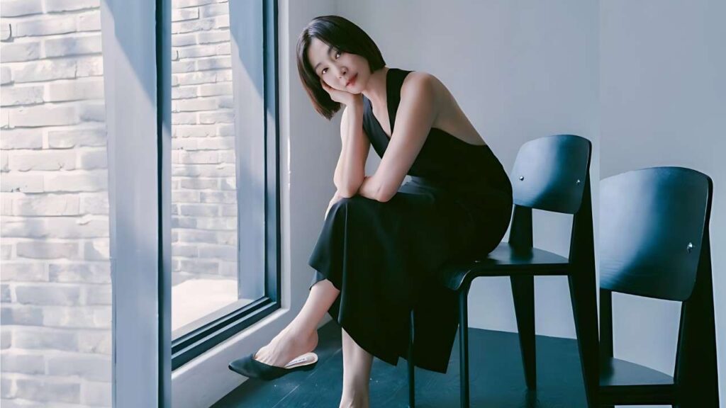 Park Hyo-joo in a sleeveless black dress sits on a chair by a large window, resting her head on her hand with flats on the floor nearby.