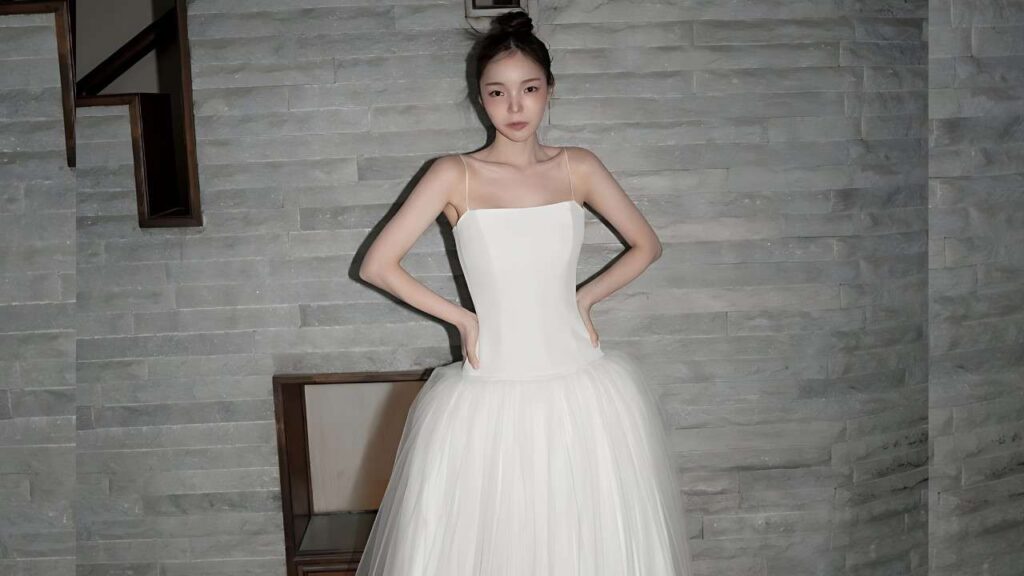 Park Jin-joo (박진주) in a white strap-shoulder wedding gown with a fitted bodice and full tulle skirt stands with hands on hips against a gray stone wall, staircase visible on the left.