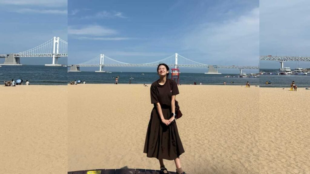 Park Ju-hyun in a dark midi dress standing center, hands clasped, as a suspension bridge spans the blue sea.