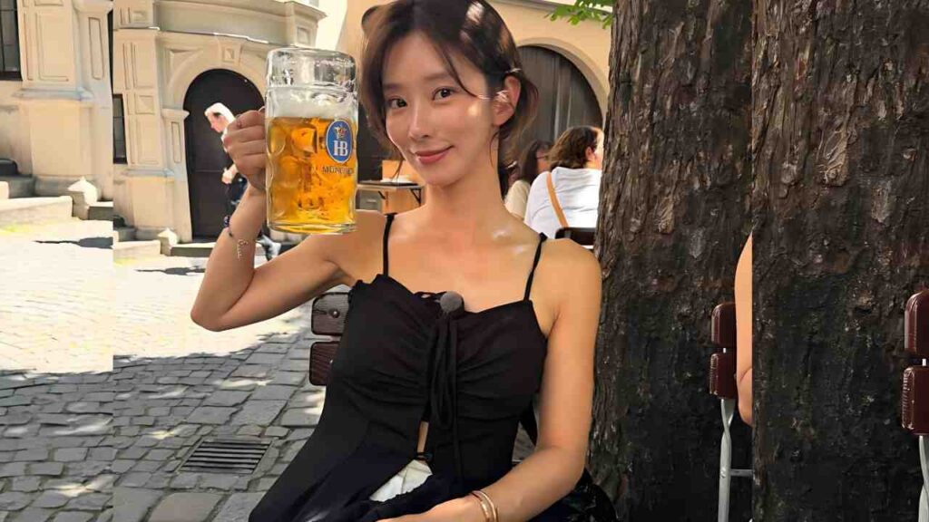 Ra Mi-ran in a black spaghetti-strap top raises a tall beer stein outdoors, beside a tree on a cobblestone square.