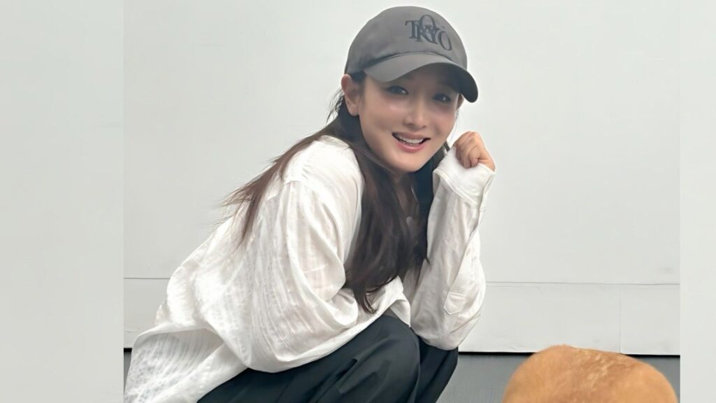 Sa Kang (사강) crouches in a loose white shirt and dark pants, smiling under a gray cap, with a small brown dog near a pale wall.