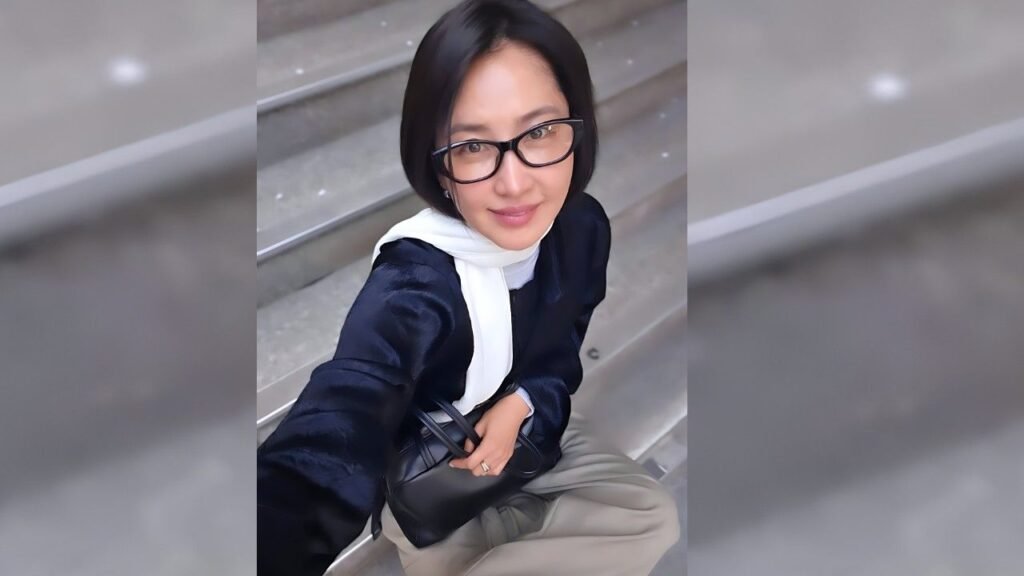 Shin Dong-mi with short black hair and glasses sits on steps, wearing a navy blouse and white scarf, smiling at the camera.