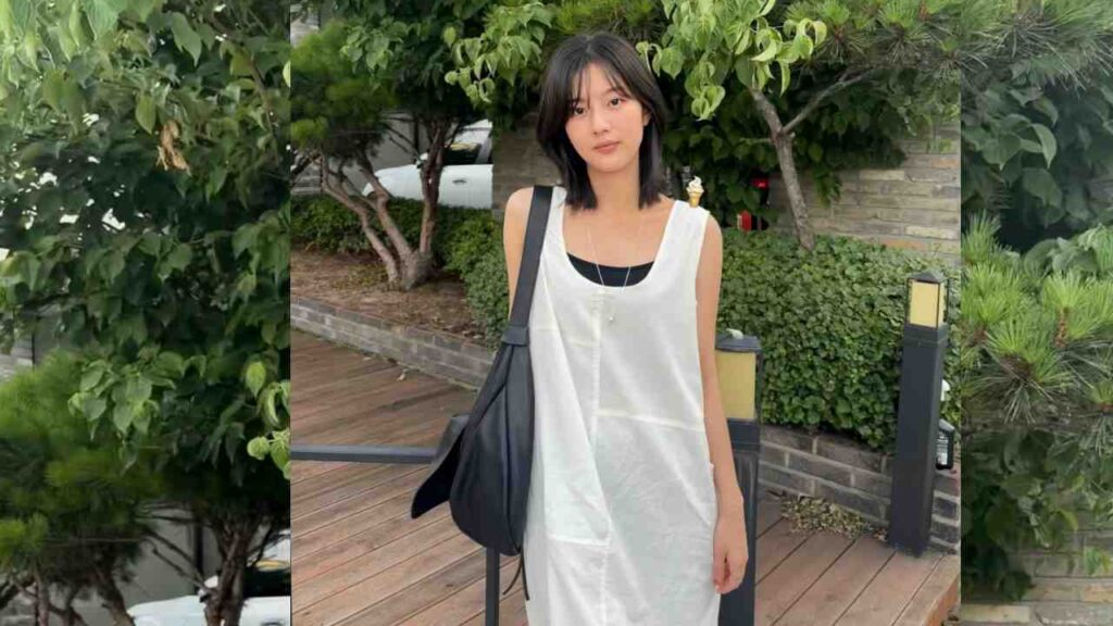 Shin Eun-soo with black hair in a white sleeveless dress over a black top, carrying a black bag, on a deck amid greenery.