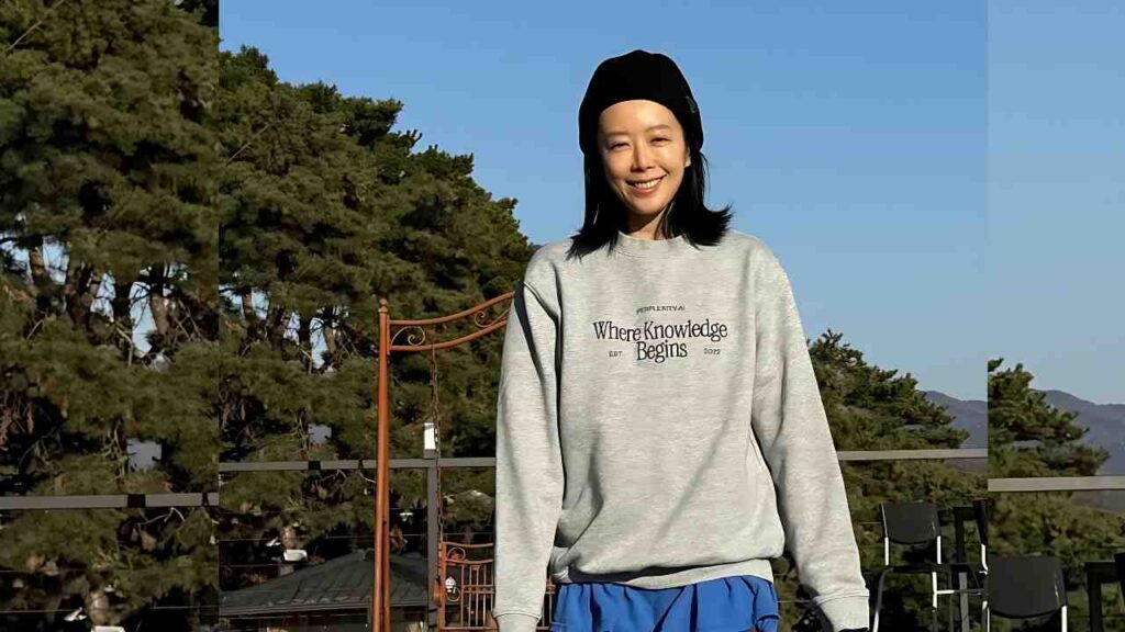 Shin Ji-soo in a gray sweatshirt reading “Where Knowledge Begins” and a black beanie, standing on an outdoor deck with trees and blue sky behind.