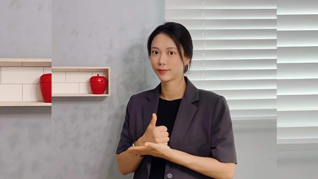 Shin Joo-ah in a dark blazer and black top poses indoors, giving a thumbs-up; pale patterned wall, red apples on a shelf, and blinds behind.