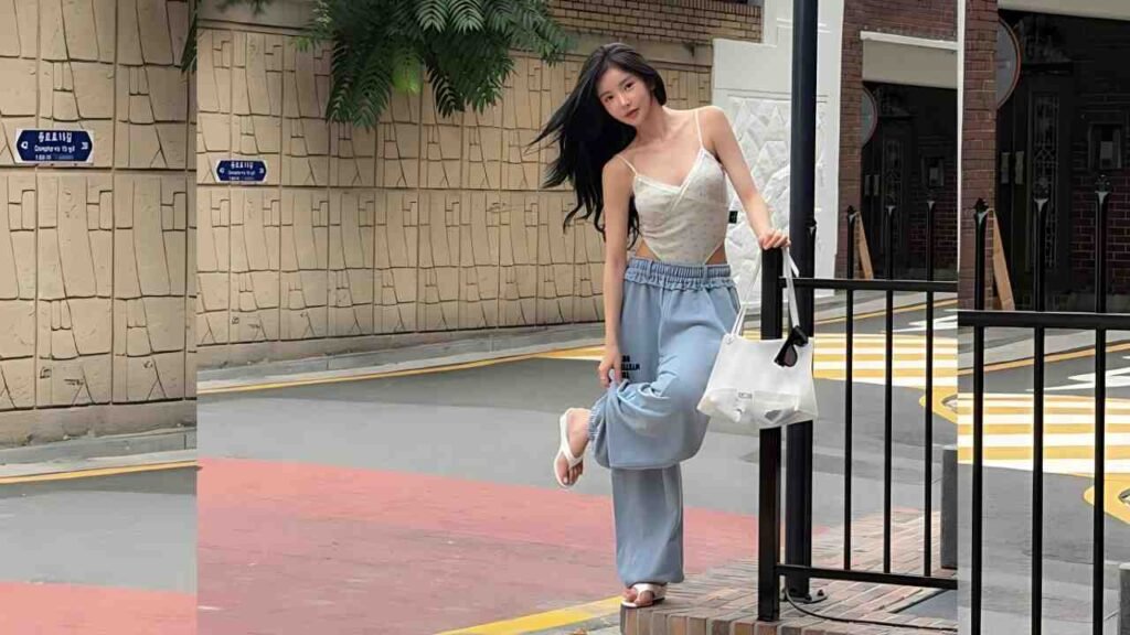Shin Sae-rom in a beige spaghetti-strap top and light blue wide-leg pants leans on a black railing, carrying a white tote on a sunlit urban street.