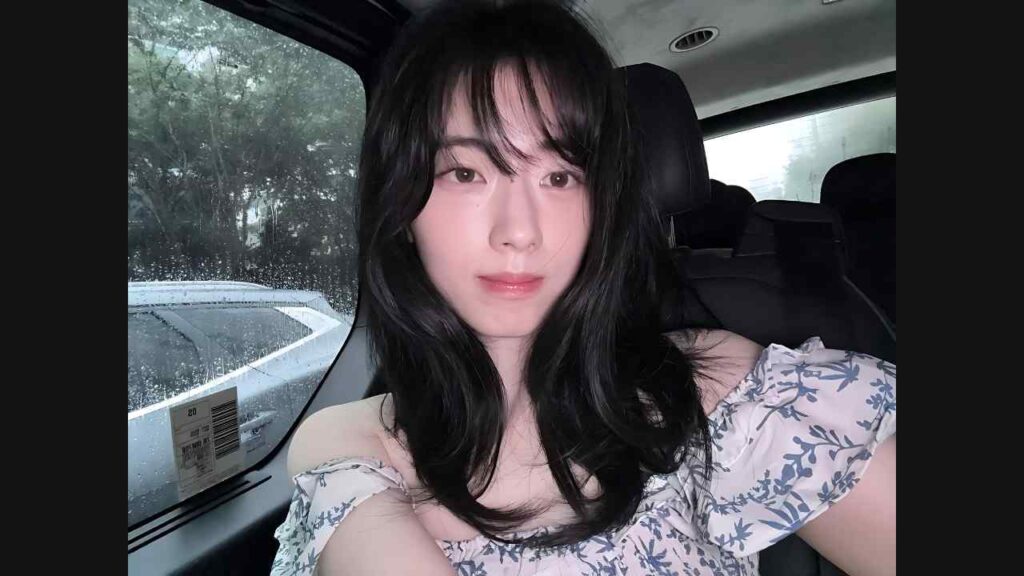 Shin Se-hwi with long dark hair and a floral off-shoulder top sits in a car, rainy window and trees outside.