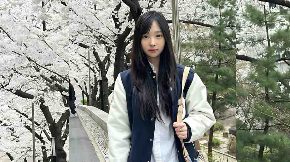 Shin Yi with long dark hair in a navy varsity jacket stands on a cherry blossom-lined bridge, beige bag over shoulder.